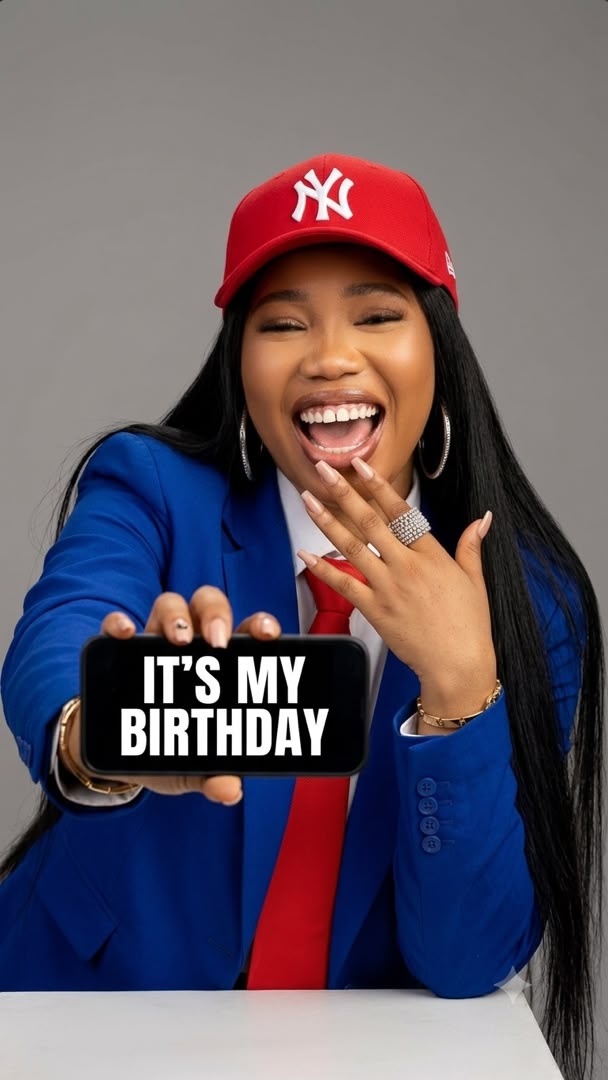 Woman in a red cap and blue blazer holding up a phone that reads It's My Birthday