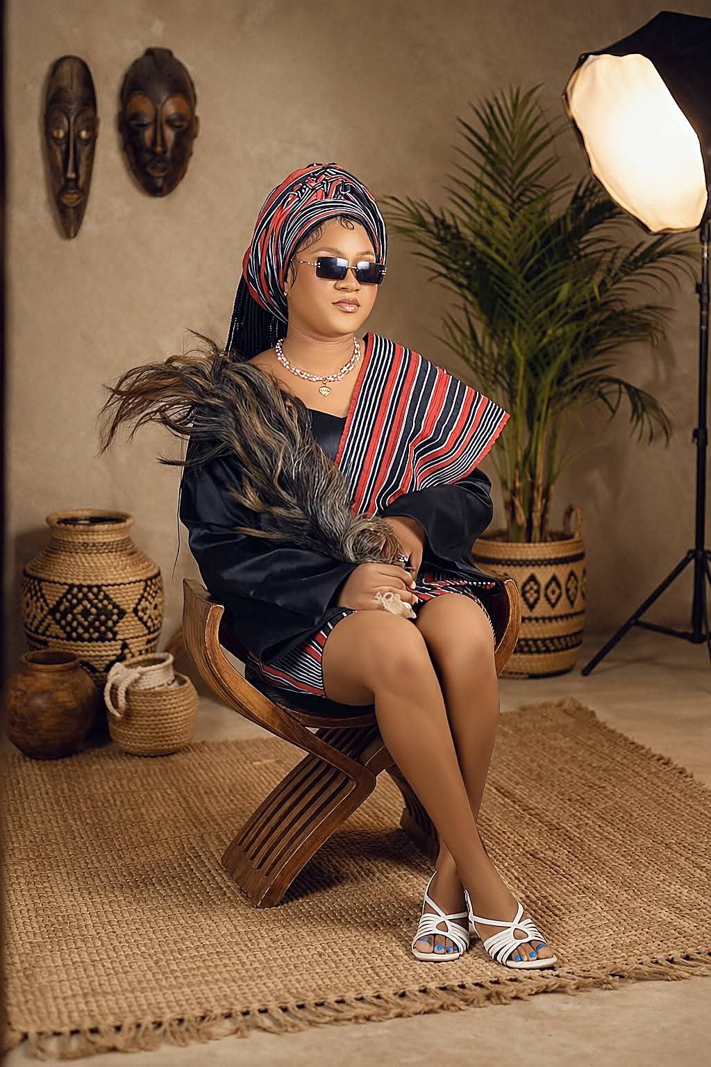 Woman in a striped headwrap and sunglasses seated on a carved wooden chair styled with African masks and woven baskets