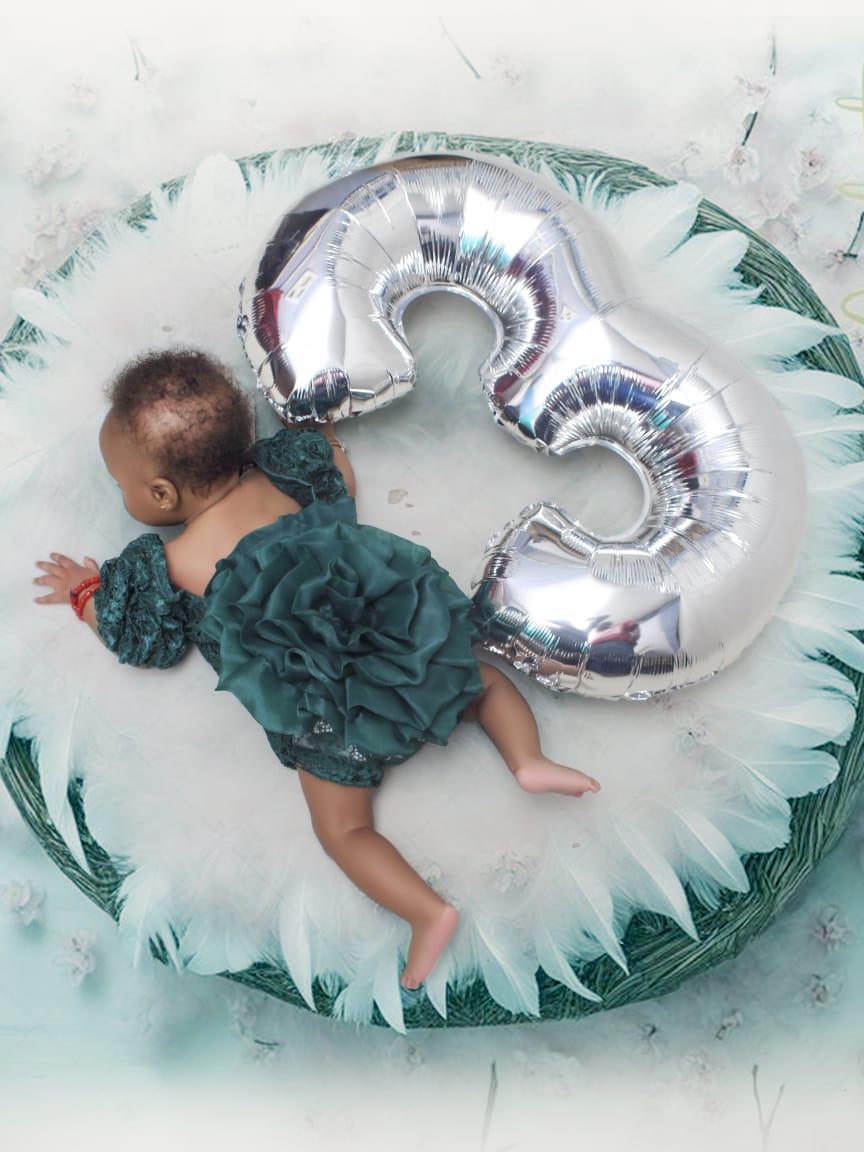 Baby lying beside a giant silver number three balloon on a feathered set
